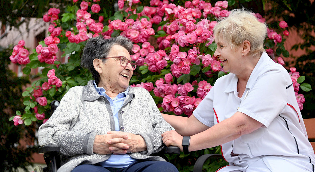 Zwei Personen unterhalten sich und lachen freundlich, sie sitzen vor einem Busch und zahlreichen rosa Blüten.