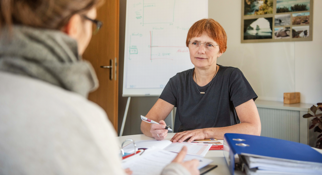 Zwei Personen sitzen in Beratungssituation gegenüber, machen Notizen. Im Hintergrund ein Flipchart.