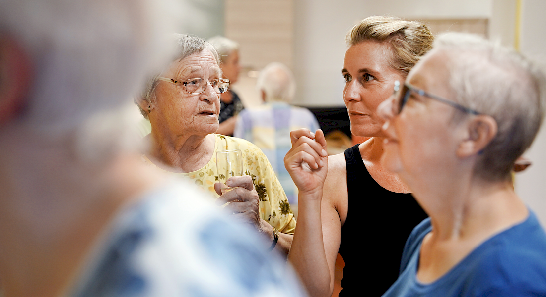 Drei Personen bei Veranstaltung im Bild, im Hintergrund im Bildfokus zwei Personen im intensiven Gespräch.