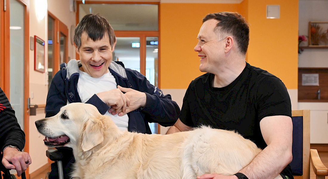 Zwei Personen sitzen mit einem Hund im Raum und strahlen.