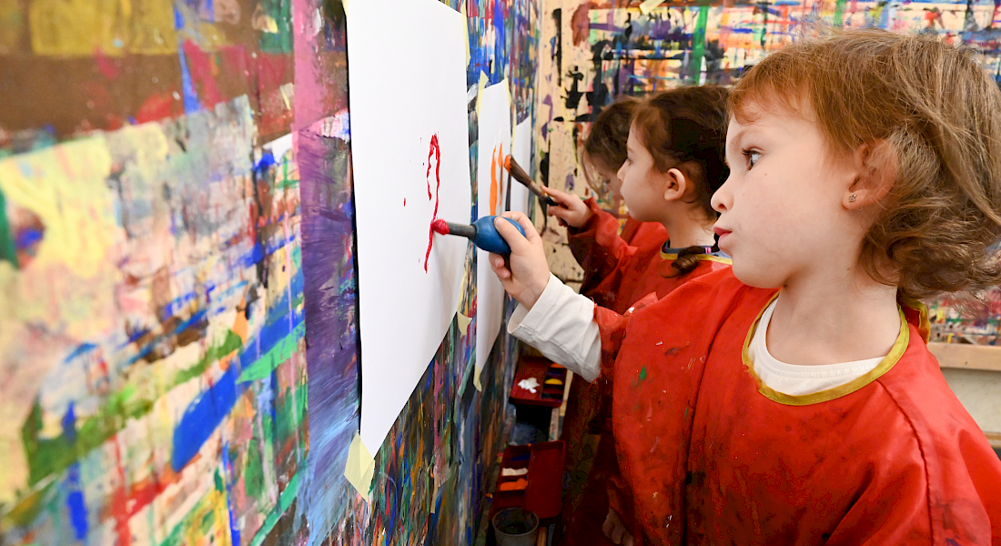 Kita-Kinder malen mit Farben Bilder, die an der Wand hängen. Die Wand ist übersät mit Farbtupfern.