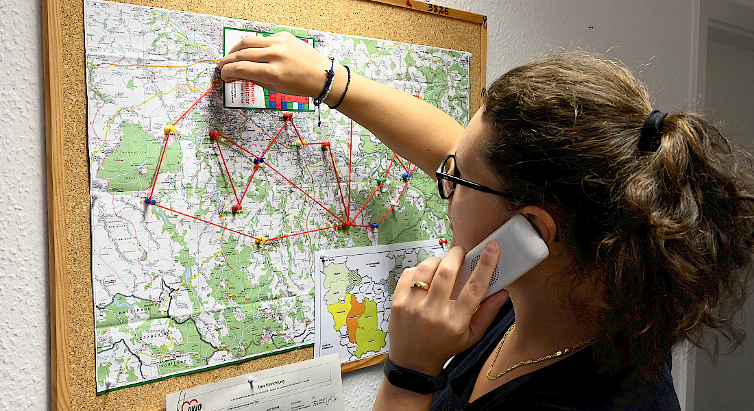 Person steht mit Telefon am Ohr an einer Pinnwand, dort ist eine Landkarte angepinnt.