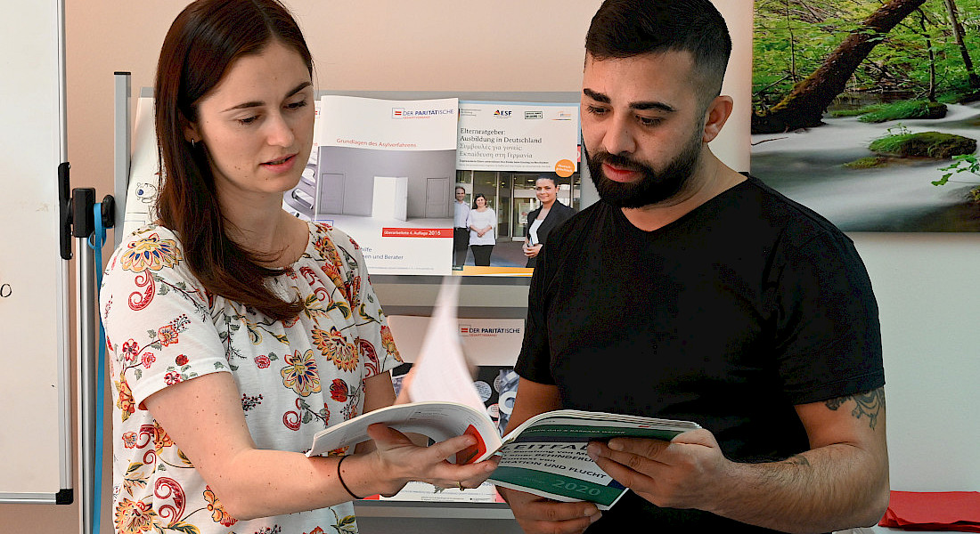 Zwei Personen stehen vor einem Flyerständer, blättern ein Heft durch.